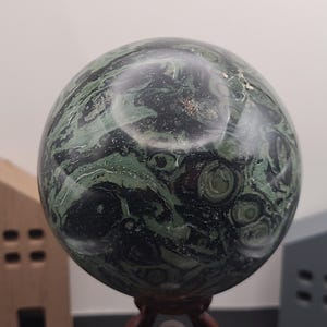 May include: A green and black sphere with swirling patterns, made of polished stone. The sphere is on a wooden stand.