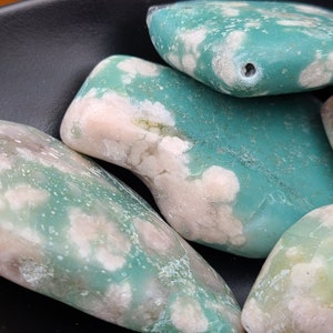 May include: Close-up of three smooth, polished stones with a mottled green and white pattern. The stones have a natural, organic shape and a glossy finish.