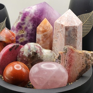 May include: A collection of polished pink, red, and purple crystals arranged in a circular wooden bowl. The crystals include a heart-shaped pink crystal, a large purple crystal, and several smaller crystals.