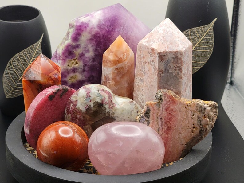 May include: A collection of polished pink, red, and purple crystals arranged in a circular wooden bowl. The crystals include a heart-shaped pink crystal, a large purple crystal, and several smaller crystals.