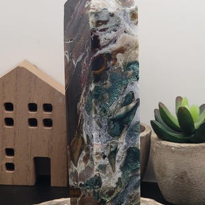 May include: A green, brown, and white agate crystal point with a polished finish. The crystal is sitting on a wooden base.