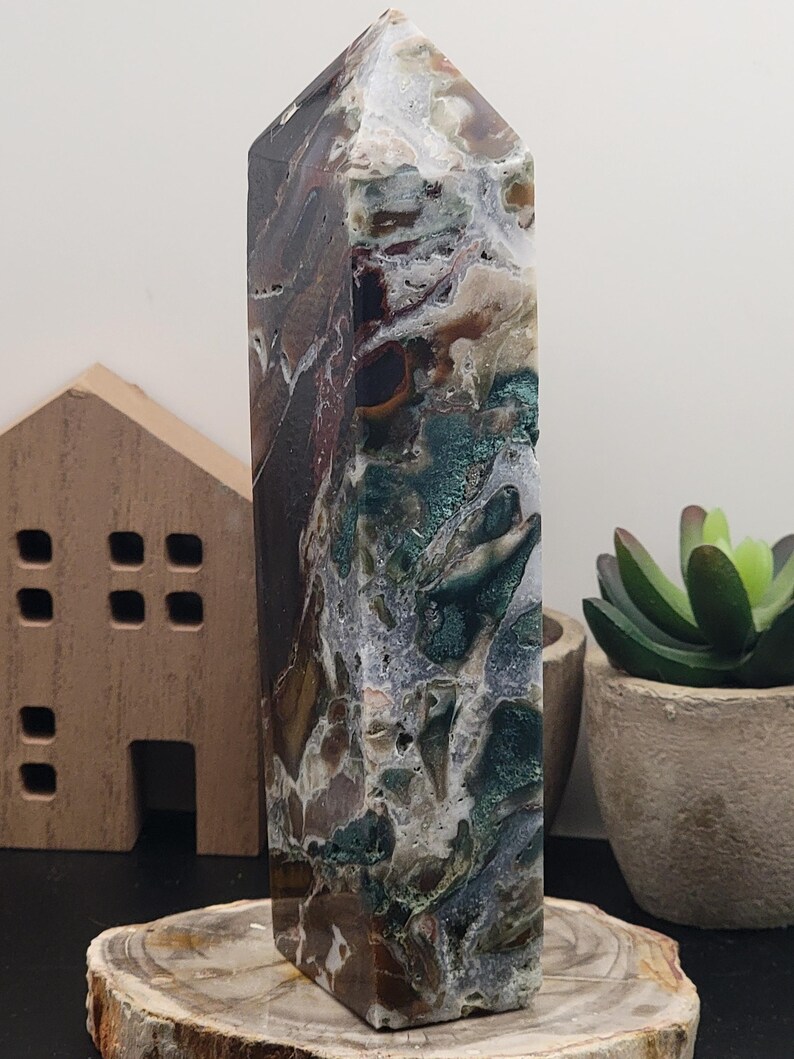 May include: A green, brown, and white agate crystal point with a polished finish. The crystal is sitting on a wooden base.