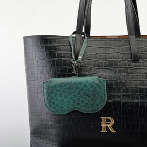 May include: A black leather tote bag with a crocodile texture, featuring a gold-colored letter "R" emblem. A green leather glasses case is attached to the bag with a silver-colored clasp. The bag has black handles.