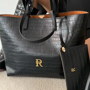 Puede incluir: Un bolso tote negro con textura de cocodrilo, que cuenta con dos asas superiores y un logotipo de la letra R en tono dorado en la parte frontal. Un estuche con cremallera a juego, también con relieve de cocodrilo y logo R dorado, está unido.