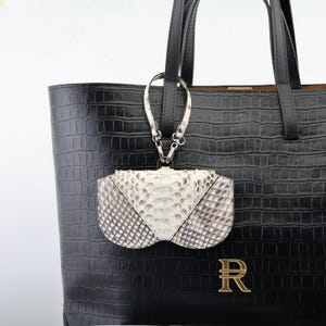 May include: A black leather tote bag with a textured crocodile pattern. A small, white and gray snakeskin clutch hangs from the handle. The bag features a gold-colored letter "R" on the front.
