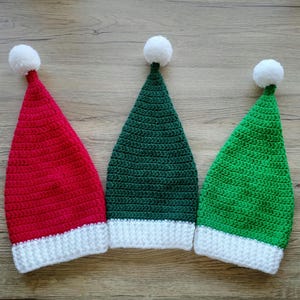 Peut inclure: Trois bonnets de No&euml;l crochet&eacute;s, rouge, vert et vert fonc&eacute;, chacun avec un bord blanc et un pompon blanc sur le dessus. Les chapeaux sont dispos&eacute;s c&ocirc;te &agrave; c&ocirc;te sur une surface en bois.