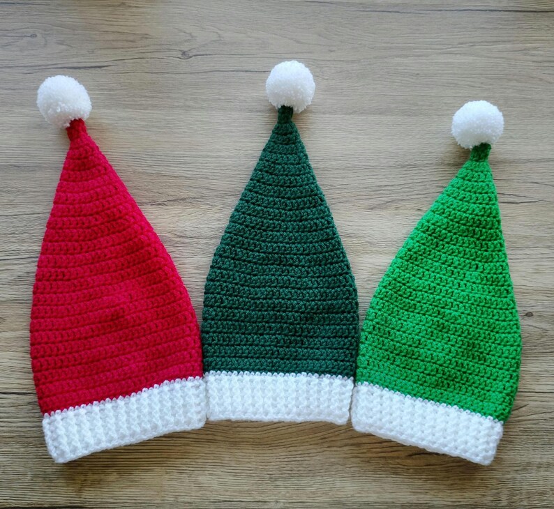 Peut inclure: Trois bonnets de No&euml;l crochet&eacute;s, rouge, vert et vert fonc&eacute;, chacun avec un bord blanc et un pompon blanc sur le dessus. Les chapeaux sont dispos&eacute;s c&ocirc;te &agrave; c&ocirc;te sur une surface en bois.