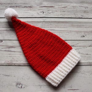 Peut inclure: Un bonnet de P&egrave;re No&euml;l rouge et blanc crochet&eacute; avec un pompon blanc au bout. Le chapeau est fait d'un fil textur&eacute; et a une bande blanche en bas. Le chapeau est pos&eacute; sur une surface en bois.