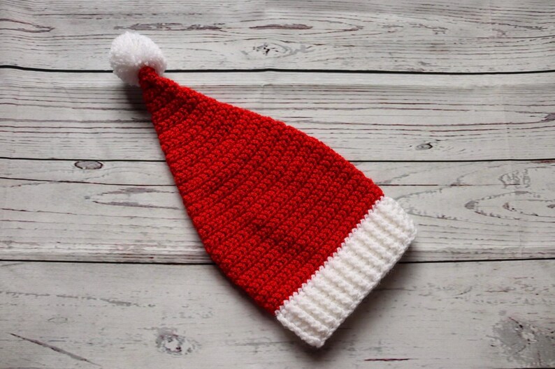 Peut inclure: Un bonnet de P&egrave;re No&euml;l rouge et blanc crochet&eacute; avec un pompon blanc au bout. Le chapeau est fait d'un fil textur&eacute; et a une bande blanche en bas. Le chapeau est pos&eacute; sur une surface en bois.