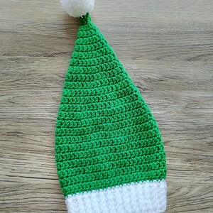 Peut inclure: Un chapeau vert crochet&eacute; &agrave; la main avec un bord blanc et un pompon blanc sur le dessus. Le chapeau est de forme conique et semble &ecirc;tre pour une occasion festive. Les points de crochet sont clairement visibles, mettant en valeur le savoir-faire.