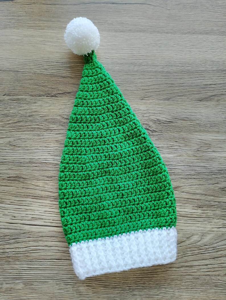 Peut inclure: Un chapeau vert crochet&eacute; &agrave; la main avec un bord blanc et un pompon blanc sur le dessus. Le chapeau est de forme conique et semble &ecirc;tre pour une occasion festive. Les points de crochet sont clairement visibles, mettant en valeur le savoir-faire.
