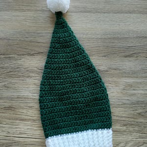 Peut inclure: Un chapeau vert en crochet fait &agrave; la main avec un bord blanc et un pompon blanc sur le dessus. Le chapeau est de forme conique et semble &ecirc;tre fait de fil doux. Le chapeau est probablement pour un enfant ou une occasion festive.