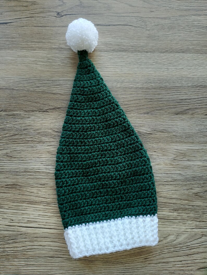 Peut inclure: Un chapeau vert en crochet fait &agrave; la main avec un bord blanc et un pompon blanc sur le dessus. Le chapeau est de forme conique et semble &ecirc;tre fait de fil doux. Le chapeau est probablement pour un enfant ou une occasion festive.