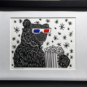 May include: A black and white illustration of a bear wearing 3D glasses and holding a bucket of popcorn. The background is white with black stars. The text "Movie Night" is at the bottom of the image.