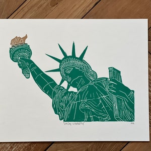 May include: A green and tan linocut print on white paper depicting the Statue of Liberty holding a torch and a tablet. The text "LADY LIBERTY" is printed in black ink at the bottom center, with the initials "AK" in the bottom right corner.