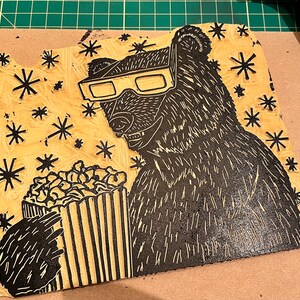 May include: A black and white woodblock print of a bear wearing 3D glasses and holding a bucket of popcorn. The background is a light brown with a pattern of black stars.