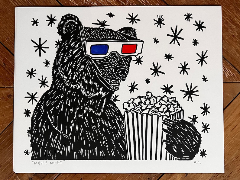 May include: A black and white linocut print of a bear wearing 3D glasses and holding a bucket of popcorn. The bear is surrounded by small black stars. The print is titled "Movie Night" and signed "AK".
