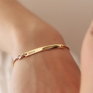 May include: A gold bracelet with a rectangular nameplate engraved with the name "Jessica". The bracelet has a delicate chain and small, pink gemstones. The jewellery is worn on a wrist.