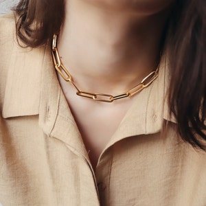 以下が含まれることがあります： 大きな長方形のリンクを持つゴールドチェーンネックレス。ネックレスは、長い茶色の髪をした人が着用しています。