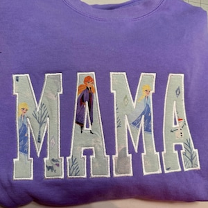 May include: A lavender crewneck sweatshirt featuring the word MAMA in large block letters. The letters are filled with a light blue fabric pattern depicting characters and winter scenery from the movie Frozen, outlined in white embroidery thread.