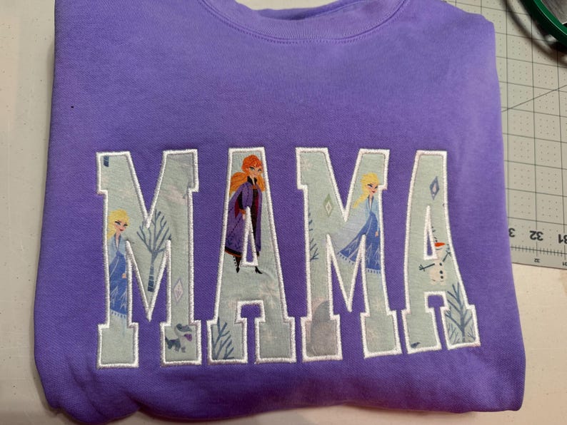 May include: A lavender crewneck sweatshirt featuring the word MAMA in large block letters. The letters are filled with a light blue fabric pattern depicting characters and winter scenery from the movie Frozen, outlined in white embroidery thread.