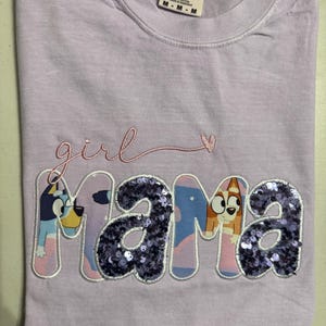 May include: A light purple short-sleeve t-shirt with a crew neckline. The shirt features the embroidered word girl in pink script above the word mama. The letters in mama are filled with purple sequins and cartoon dog characters.