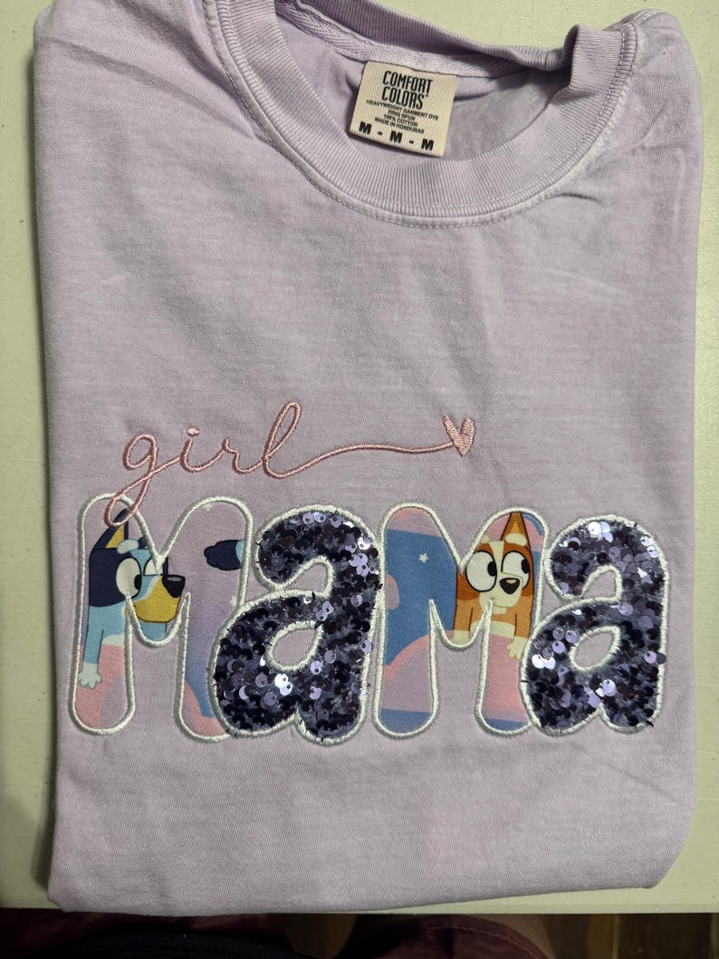 May include: A light purple short-sleeve t-shirt with a crew neckline. The shirt features the embroidered word girl in pink script above the word mama. The letters in mama are filled with purple sequins and cartoon dog characters.