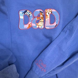 May include: A periwinkle blue long-sleeve sweatshirt featuring the word DAD embroidered in large letters filled with a cartoon character pattern and outlined in pink. The sleeve cuff is embroidered with the names Cash and Camryn in pink thread.