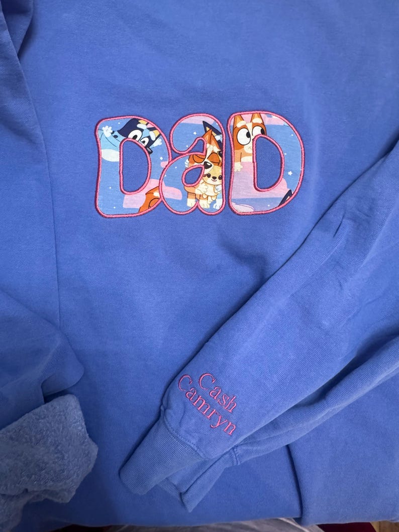 May include: A periwinkle blue long-sleeve sweatshirt featuring the word DAD embroidered in large letters filled with a cartoon character pattern and outlined in pink. The sleeve cuff is embroidered with the names Cash and Camryn in pink thread.