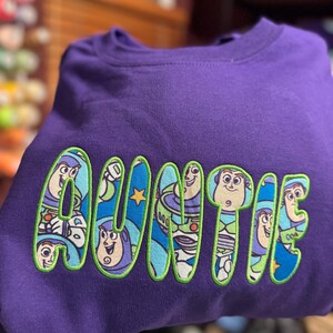 Puede incluir: Una sudadera morada de cuello redondo con la palabra AUNTIE bordada en letras grandes y llamativas. Cada letra est&aacute; rellena con un patr&oacute;n de tela azul que muestra al personaje Buzz Lightyear y delineada con costuras verde brillante.