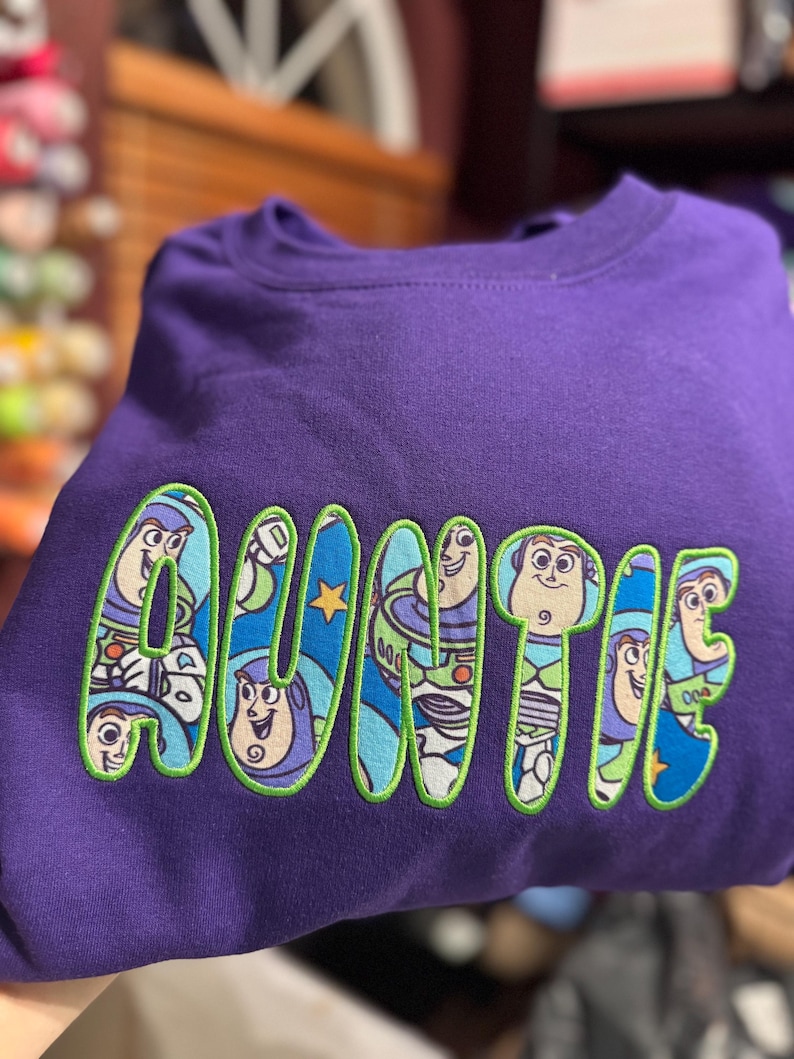 Puede incluir: Una sudadera morada de cuello redondo con la palabra AUNTIE bordada en letras grandes y llamativas. Cada letra est&aacute; rellena con un patr&oacute;n de tela azul que muestra al personaje Buzz Lightyear y delineada con costuras verde brillante.