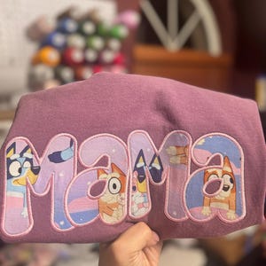 May include: A mauve-colored t-shirt featuring the word MAMA in large, block-style letters. The letters are filled with a patterned fabric depicting cartoon dog characters in shades of blue, orange, and white, outlined with pink embroidery stitching.