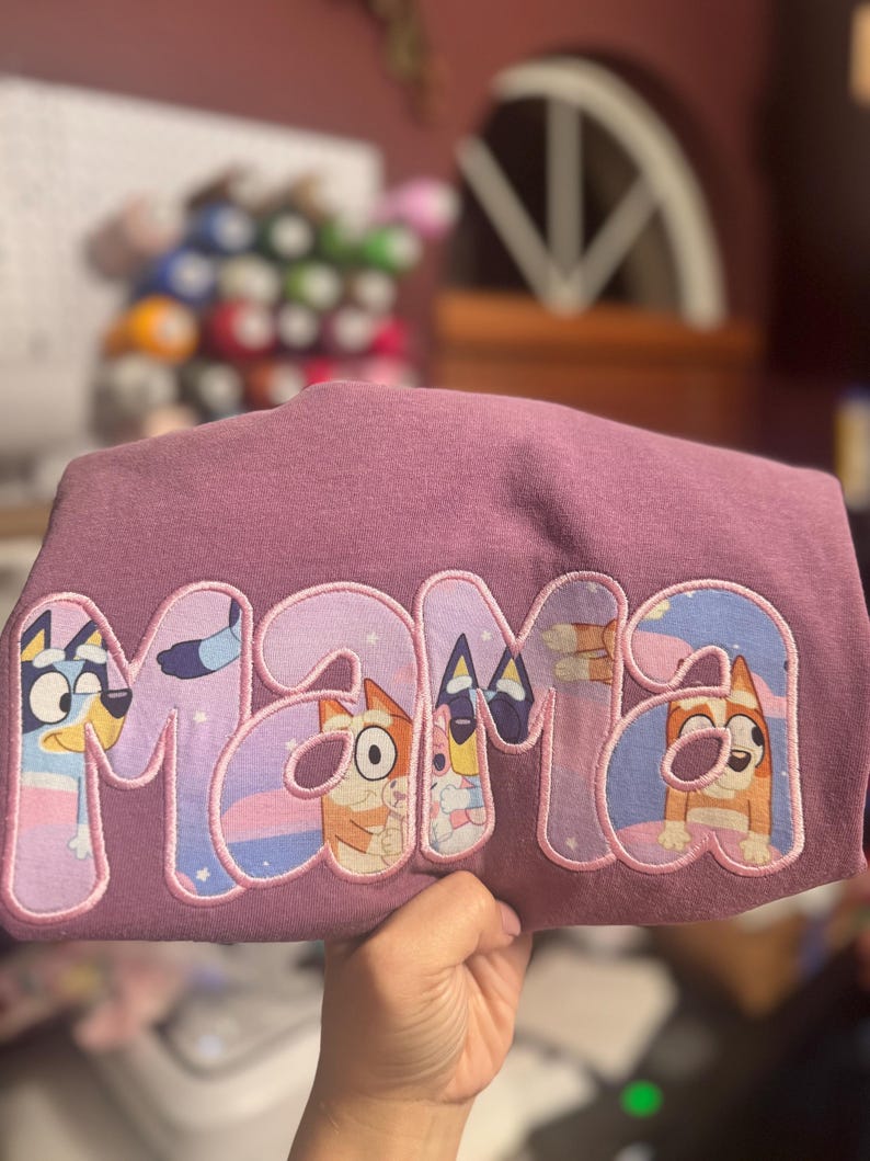 May include: A mauve-colored t-shirt featuring the word MAMA in large, block-style letters. The letters are filled with a patterned fabric depicting cartoon dog characters in shades of blue, orange, and white, outlined with pink embroidery stitching.