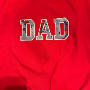 K&ouml;nnte beinhalten: Ein leuchtend rotes Sweatshirt mit Rundhalsausschnitt und dem Wort DAD als Applikation auf der Brust. Die Buchstaben sind mit einem grauen Stoff gef&uuml;llt, der Spider-Man in verschiedenen Posen vor einer Skyline und Spinnennetzen zeigt.