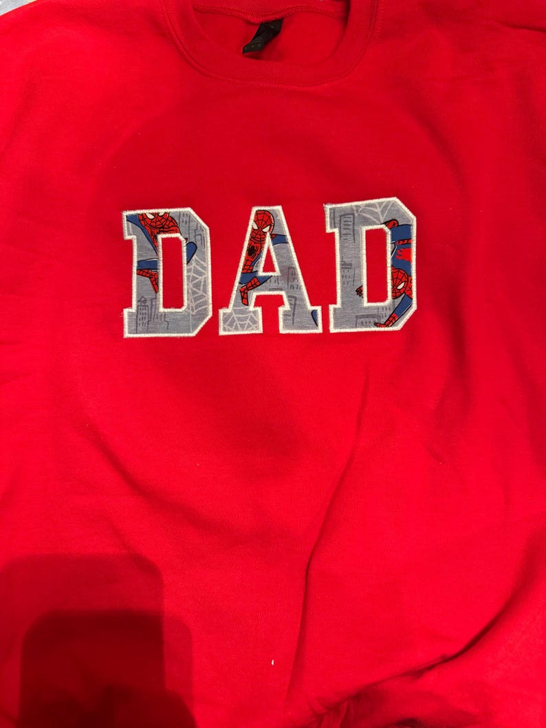 K&ouml;nnte beinhalten: Ein leuchtend rotes Sweatshirt mit Rundhalsausschnitt und dem Wort DAD als Applikation auf der Brust. Die Buchstaben sind mit einem grauen Stoff gef&uuml;llt, der Spider-Man in verschiedenen Posen vor einer Skyline und Spinnennetzen zeigt.