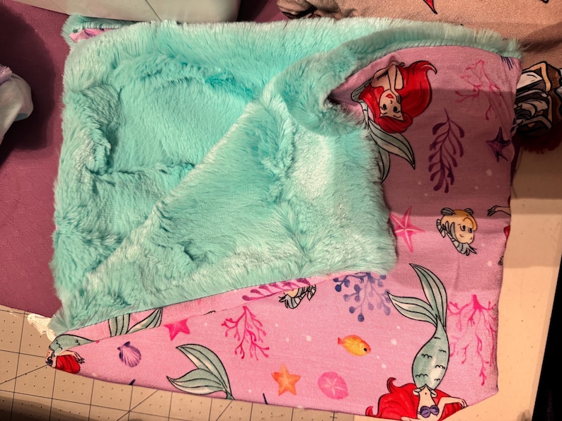 Puede incluir: Una manta de felpa turquesa con una textura suave y peluda. La parte inferior presenta una tela rosa con un tema de sirenas, que incluye ilustraciones de sirenas, peces y corales. La manta est&aacute; doblada, mostrando ambos lados.