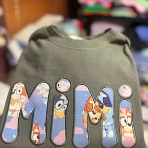May include: A dark olive green crewneck sweatshirt featuring the word "MIMI" in large, bold, embroidered letters. The letters are filled with a colorful cartoon pattern depicting blue and orange dog characters against a pink and blue sky background.