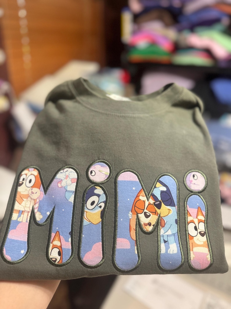 May include: A dark olive green crewneck sweatshirt featuring the word "MIMI" in large, bold, embroidered letters. The letters are filled with a colorful cartoon pattern depicting blue and orange dog characters against a pink and blue sky background.