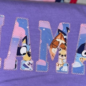 May include: A purple t-shirt featuring the word MAMA in large block letters. The letters are filled with a cartoon character pattern in shades of blue, orange, and pink, and are outlined with a decorative pink stitched border.