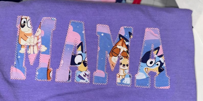 May include: A purple t-shirt featuring the word MAMA in large block letters. The letters are filled with a cartoon character pattern in shades of blue, orange, and pink, and are outlined with a decorative pink stitched border.