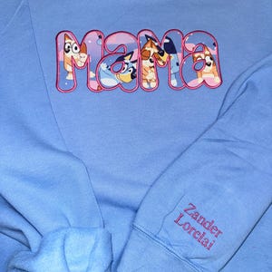 May include: A light blue sweatshirt featuring the word MAMA embroidered in pink-outlined letters filled with a cartoon dog pattern. The sleeve displays the names Zander and Lorelai embroidered in red thread on the cuff area of the garment.