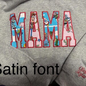 Op de afbeelding: Een grijs gem&ecirc;leerd sweatshirt met het woord MAMA in grote blokletters met een rode stikselrand. De letters zijn gevuld met een patroon van Toy Story-figuren en motieven. Op de mouw staat de geborduurde naam Oliver Skylar.