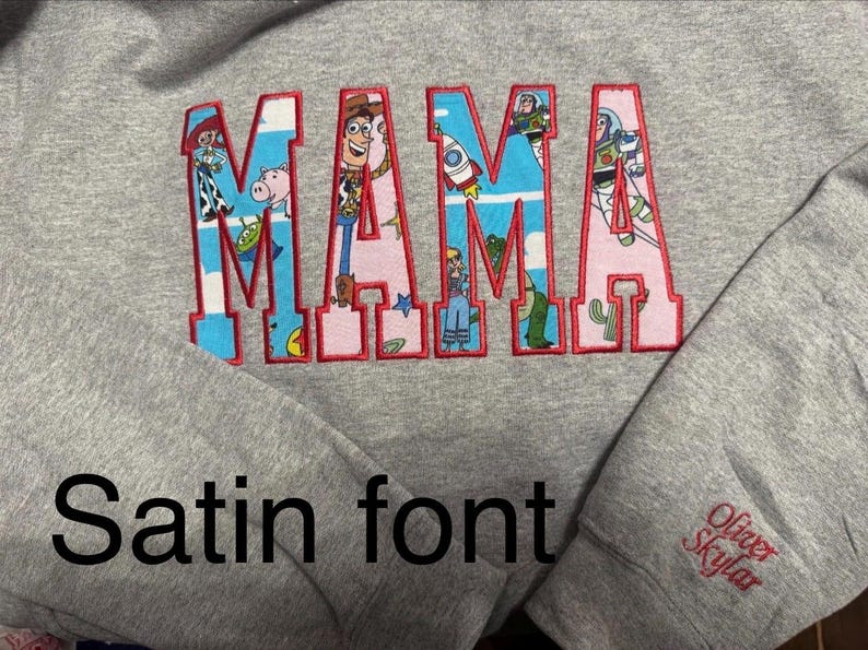 Op de afbeelding: Een grijs gem&ecirc;leerd sweatshirt met het woord MAMA in grote blokletters met een rode stikselrand. De letters zijn gevuld met een patroon van Toy Story-figuren en motieven. Op de mouw staat de geborduurde naam Oliver Skylar.