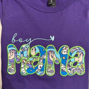 Puede incluir: Una camiseta morada de manga corta con el texto bordado 'boy mama' en letra cursiva azul claro y un peque&ntilde;o coraz&oacute;n. La palabra 'mama' est&aacute; rellena con un patr&oacute;n de tela azul que muestra un personaje de guardi&aacute;n espacial y bordes verde lima.