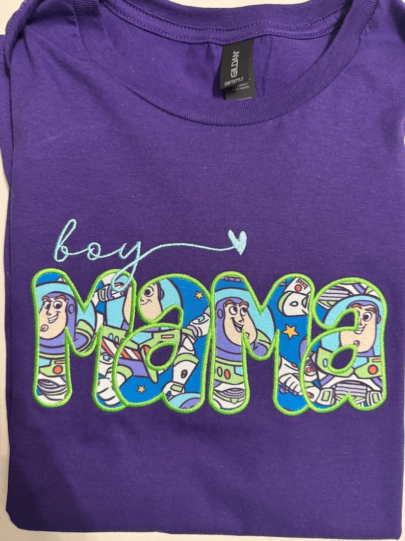 Puede incluir: Una camiseta morada de manga corta con el texto bordado 'boy mama' en letra cursiva azul claro y un peque&ntilde;o coraz&oacute;n. La palabra 'mama' est&aacute; rellena con un patr&oacute;n de tela azul que muestra un personaje de guardi&aacute;n espacial y bordes verde lima.