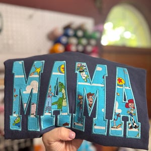 Puede incluir: Una sudadera azul marino oscuro con la palabra MAMA en letras grandes de estilo bloque. Las letras est&aacute;n rellenas con un patr&oacute;n de tela azul claro que representa varios personajes animados de Toy Story, incluidos Woody, Buzz Lightyear y Rex.
