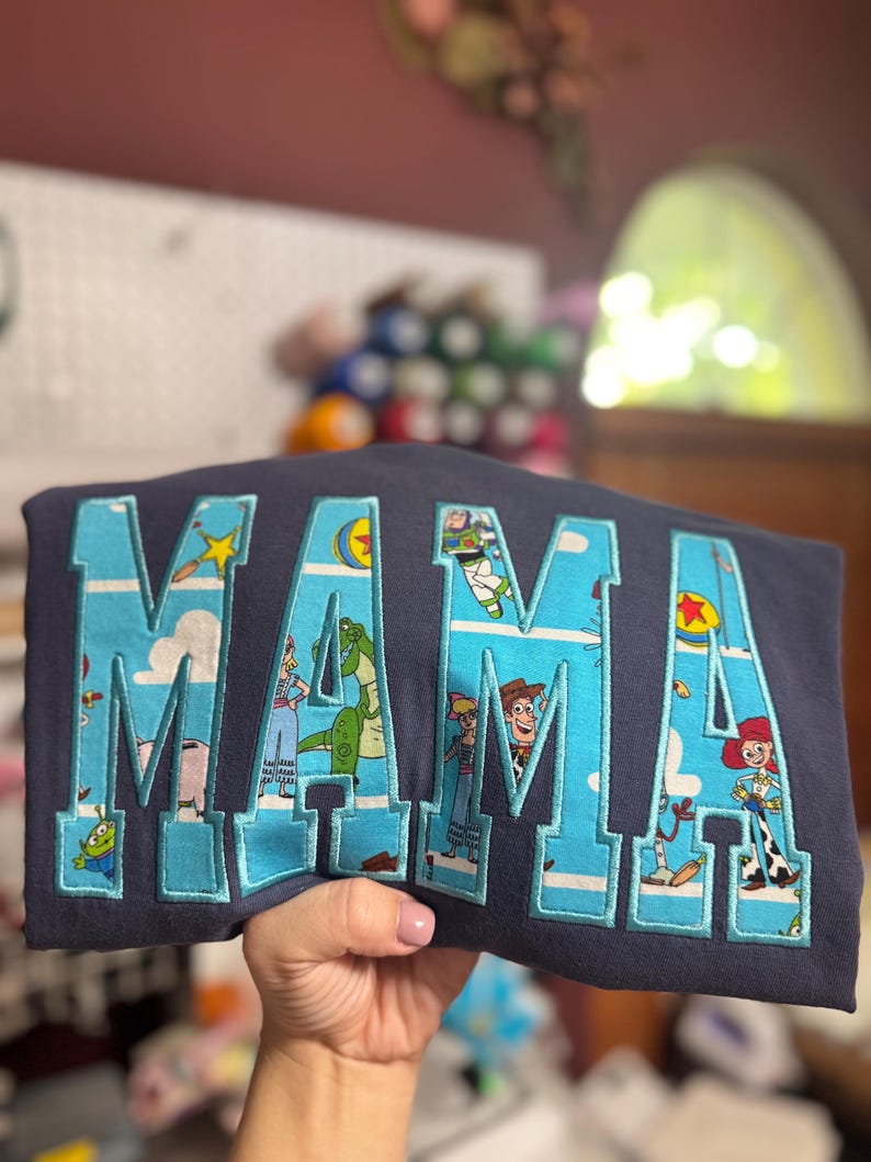 Puede incluir: Una sudadera azul marino oscuro con la palabra MAMA en letras grandes de estilo bloque. Las letras est&aacute;n rellenas con un patr&oacute;n de tela azul claro que representa varios personajes animados de Toy Story, incluidos Woody, Buzz Lightyear y Rex.