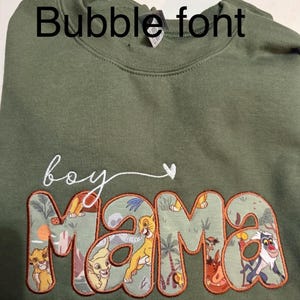 May include: An olive green crewneck sweatshirt featuring the embroidered white cursive text 'boy' followed by a small heart. Below, the word 'mama' is appliqued in a bubble font with a patterned fabric depicting various animated animal characters.