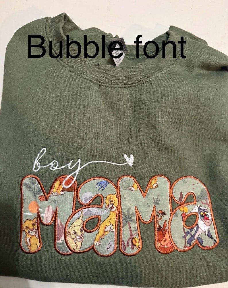 May include: An olive green crewneck sweatshirt featuring the embroidered white cursive text 'boy' followed by a small heart. Below, the word 'mama' is appliqued in a bubble font with a patterned fabric depicting various animated animal characters.