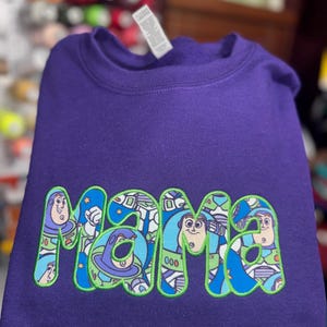 Puede incluir: Una sudadera morada de cuello redondo con la palabra "mama" bordada en una fuente de color verde brillante. El interior de cada letra est&aacute; relleno con un patr&oacute;n gr&aacute;fico colorido que representa al personaje Buzz Lightyear de Toy Story.